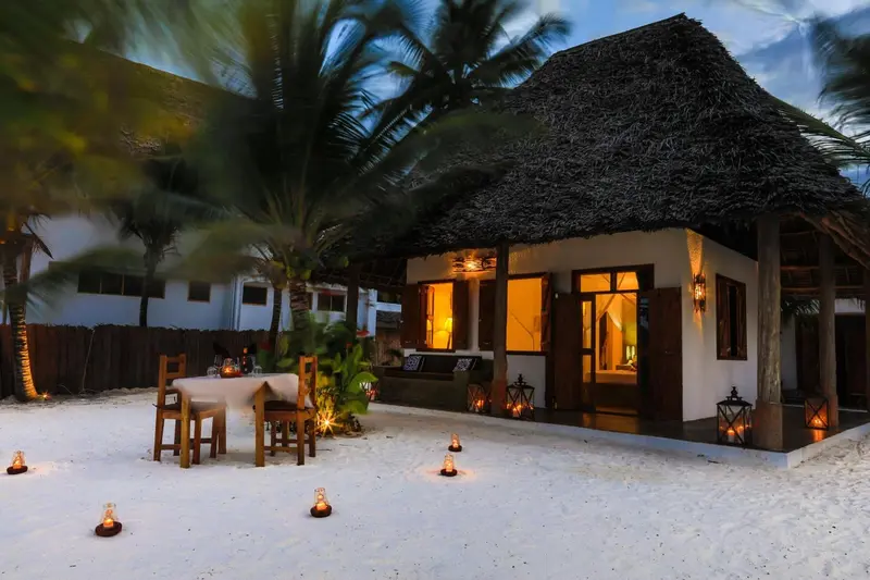 Une table et des chaises sont disposées sur le sable devant un bungalow éclairé.