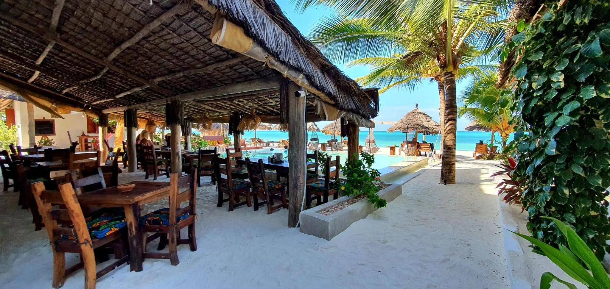 Terrasse ombragée avec tables et chaises en bois donnant sur une plage de sable blanc et la mer.