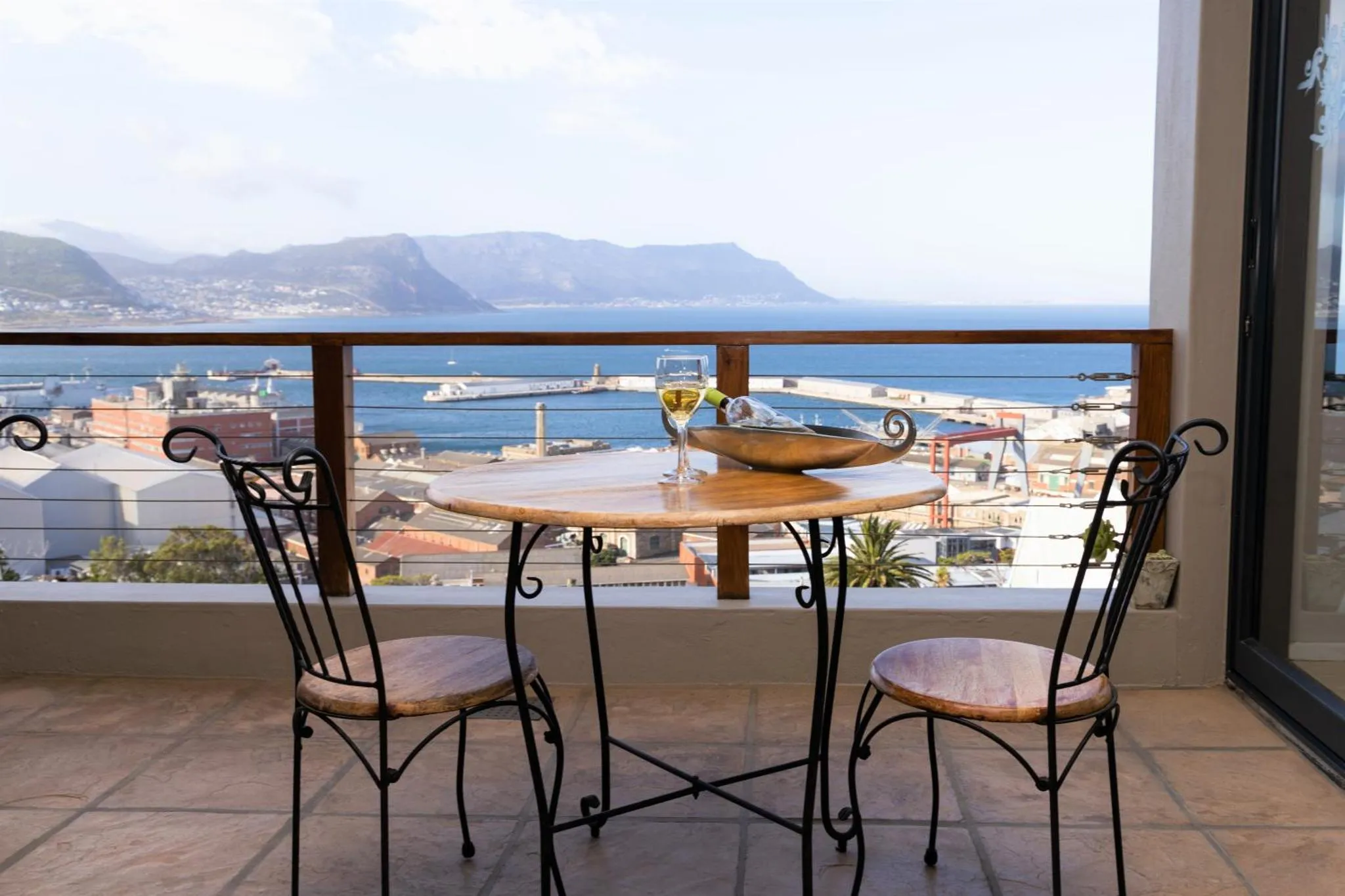 Une table ronde avec des verres et des fruits est posée sur une terrasse avec vue sur la mer et la ville.