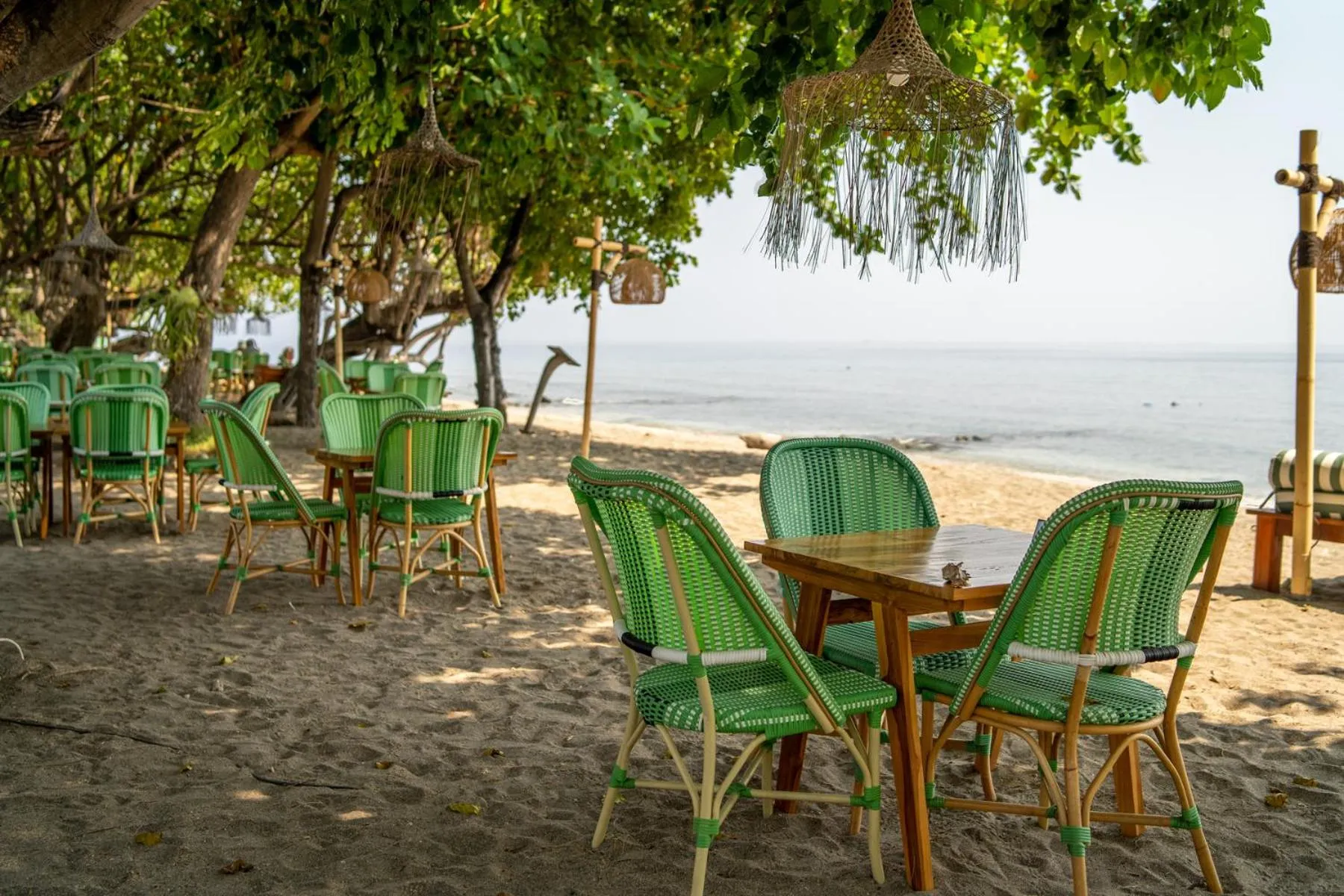 Tables et chaises vertes disposées sur une plage de sable fin, sous des arbres.