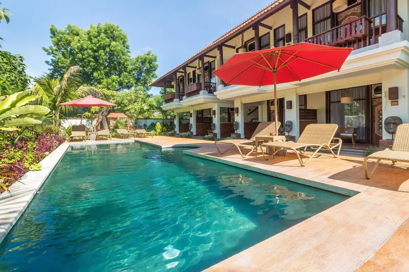 Une piscine turquoise bordée de chaises longues et de parasols rouges, devant un bâtiment à l'architecture traditionnelle.