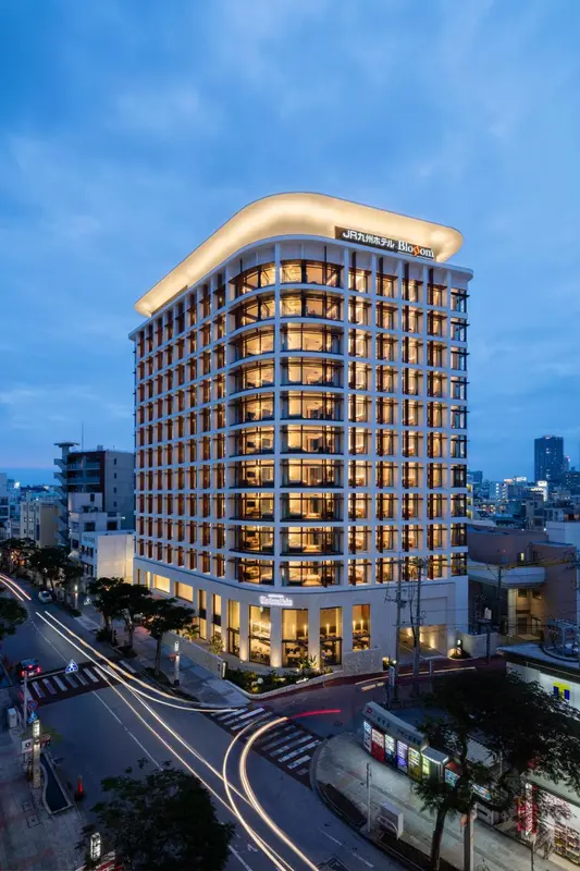 Bâtiment moderne illuminé au crépuscule avec des lumières vives dans les fenêtres et des traces de circulation en contrebas.
