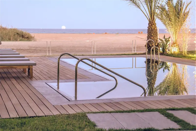 Une piscine rectangulaire bordée d'une terrasse en bois, avec une plage de sable en arrière-plan.