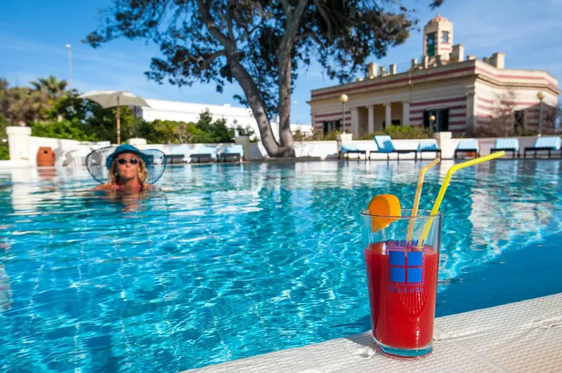 Une piscine turquoise avec une personne et un cocktail au premier plan.