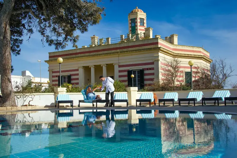 Une piscine turquoise reflète un bâtiment blanc à rayures et des transats bleus.