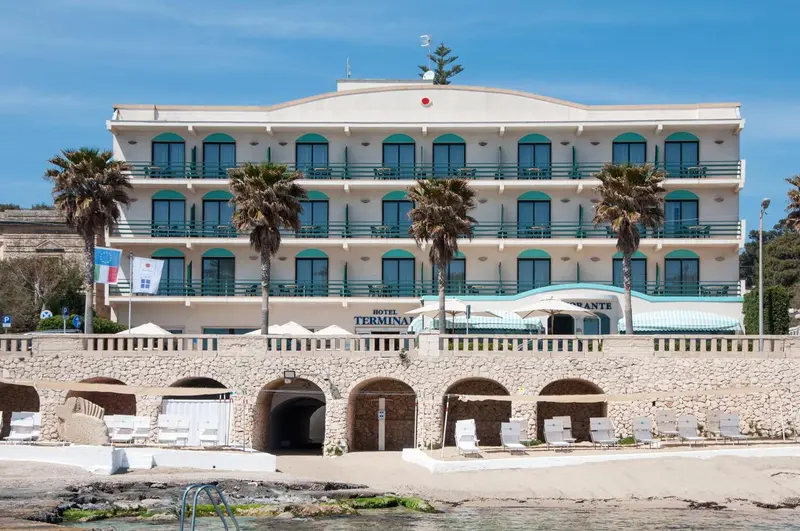 Bâtiment blanc à plusieurs étages avec des palmiers devant et une plage en contrebas.