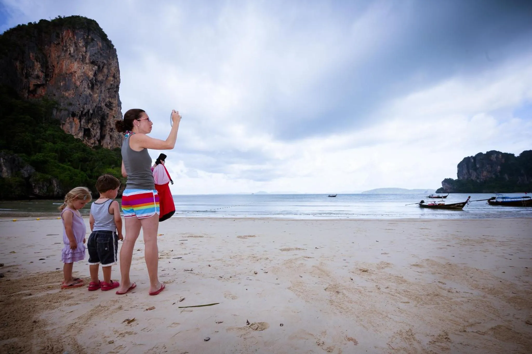 Une femme et deux enfants se tiennent sur une plage de sable fin face à la mer.