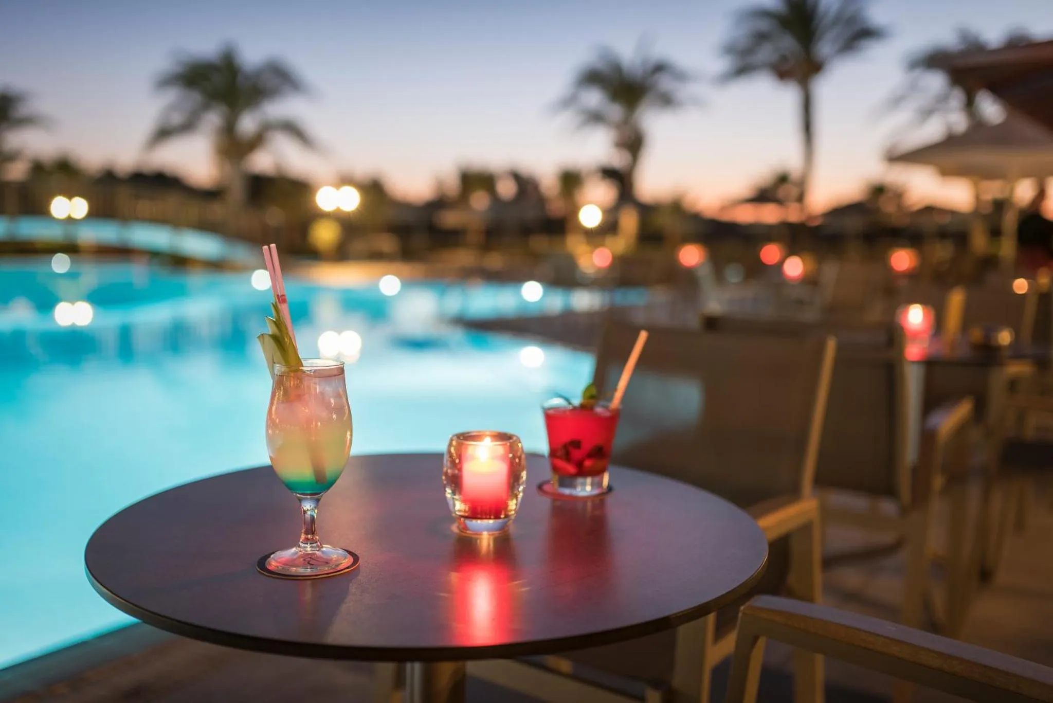 Deux cocktails colorés et une bougie illuminent une table ronde au bord d'une piscine éclairée.