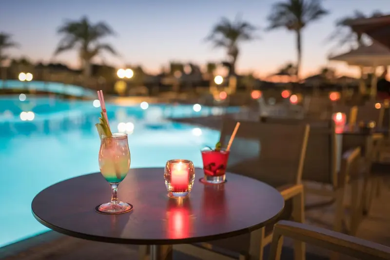 Deux cocktails colorés et une bougie illuminent une table ronde au bord d'une piscine éclairée.