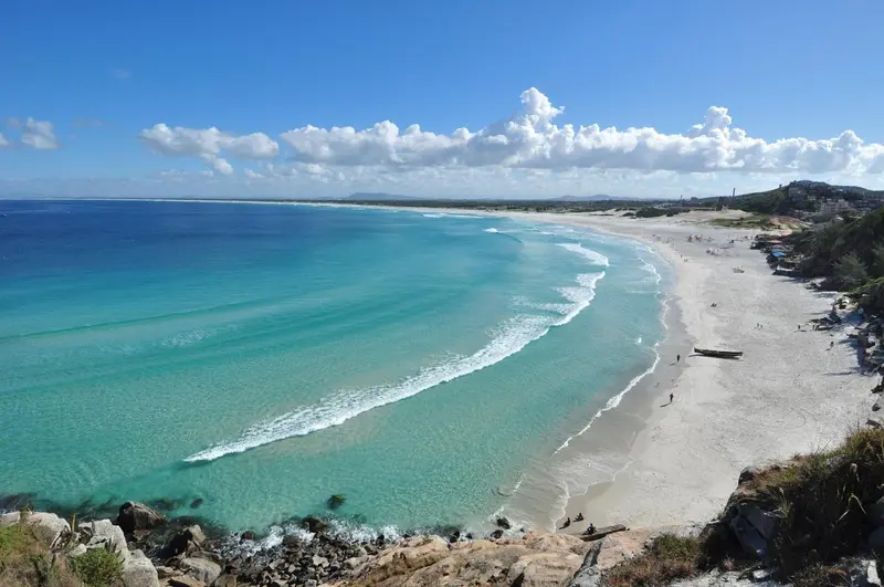 Vue aérienne d'une plage de sable fin bordée d'une mer turquoise et d'un ciel nuageux.