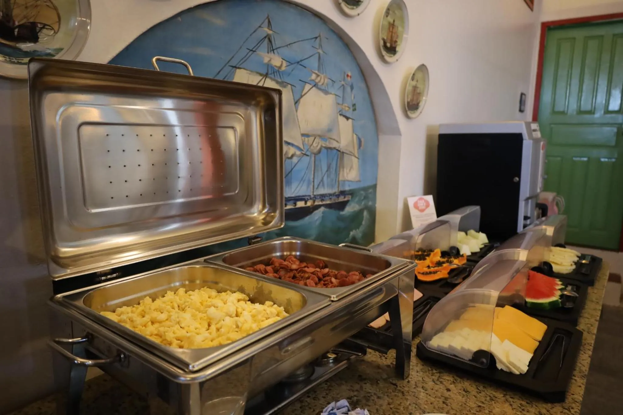 Un buffet propose des œufs brouillés, des saucisses, des fruits et du fromage.
