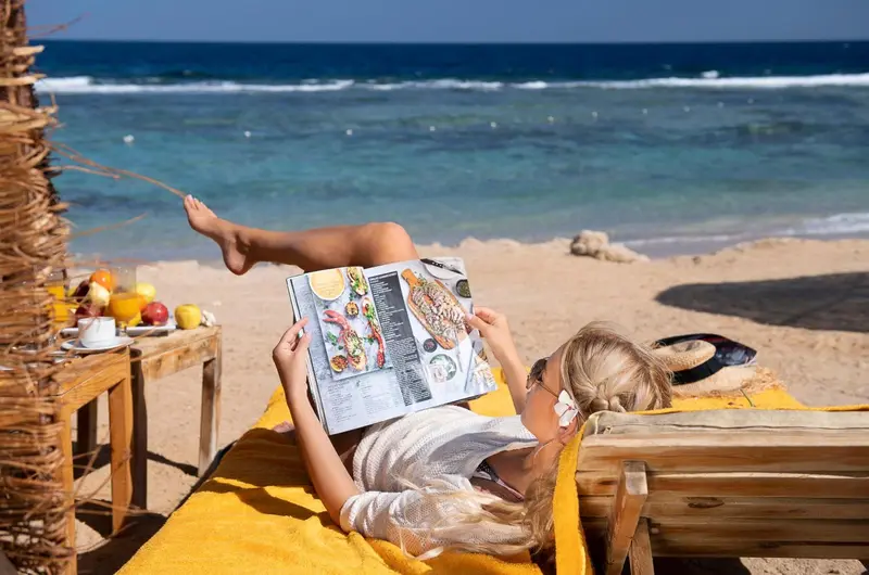 Une femme se relaxe sur une serviette jaune au bord de la mer, un livre ouvert sur les genoux.