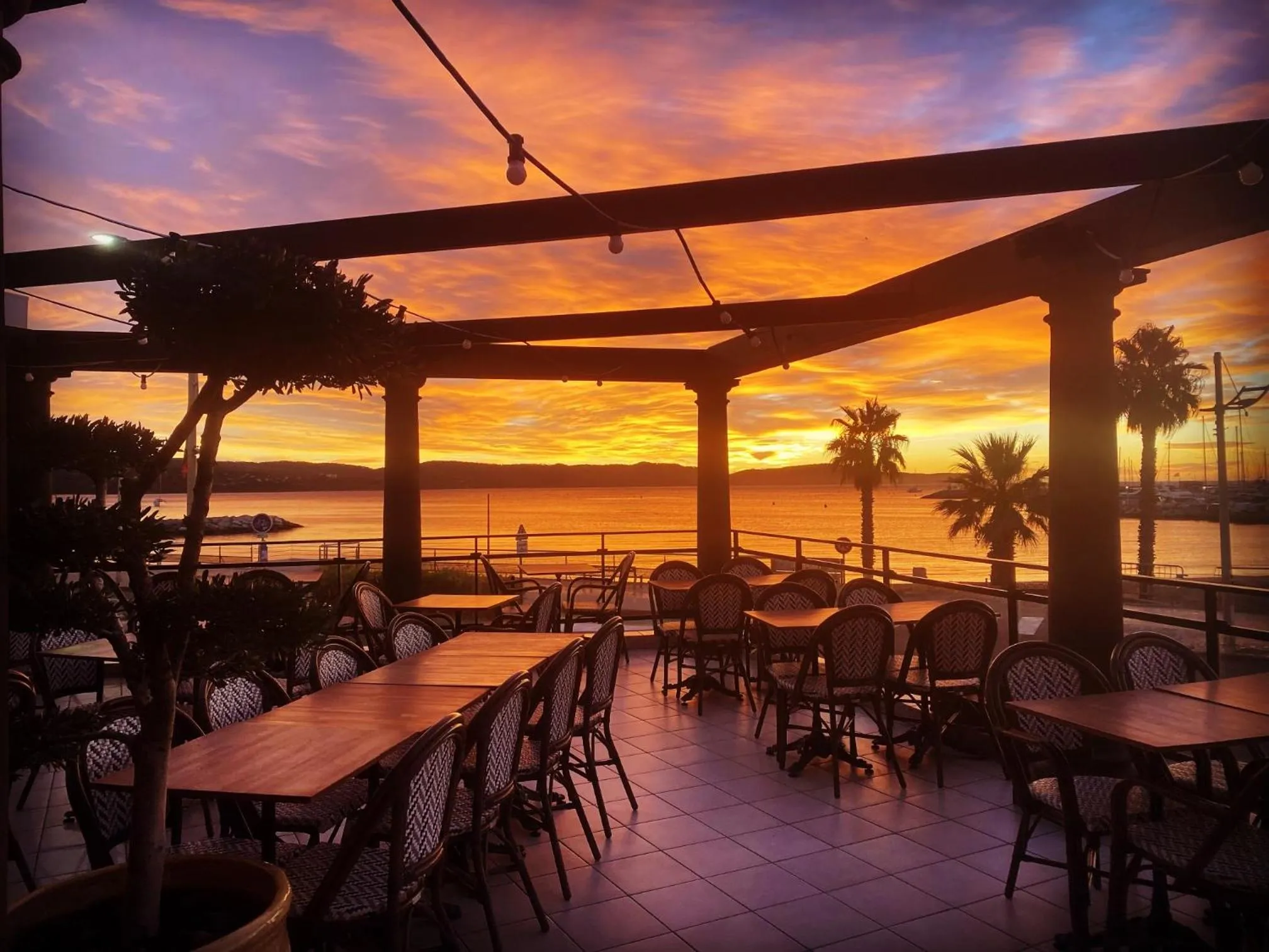 Terrasse ombragée avec tables et chaises donnant sur la mer au coucher du soleil.