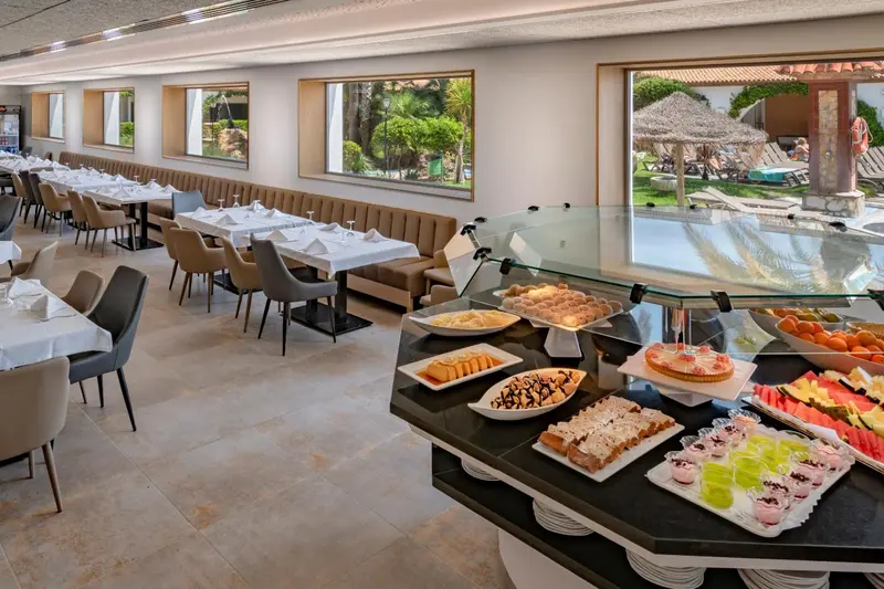 Une salle lumineuse présente un buffet garni de pâtisseries et de fruits, avec des tables dressées pour le repas.