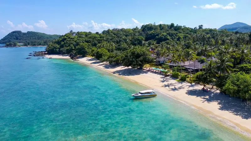 Vue aérienne d'une plage de sable fin bordée de végétation luxuriante et d'un bateau amarré dans une eau turquoise.