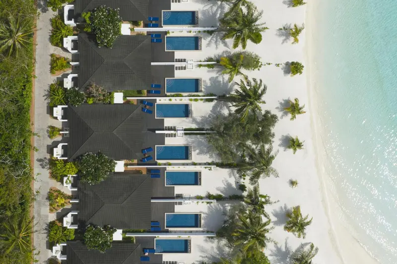 Vue aérienne de bungalows blancs avec piscines privées sur une plage de sable blanc bordée de végétation tropicale.