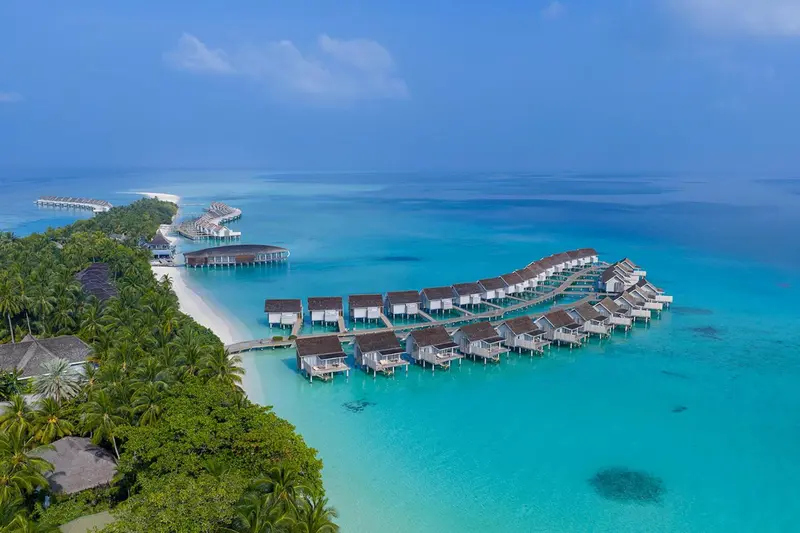 Vue aérienne d'un complexe hôtelier avec des villas sur pilotis au-dessus d'une eau turquoise.