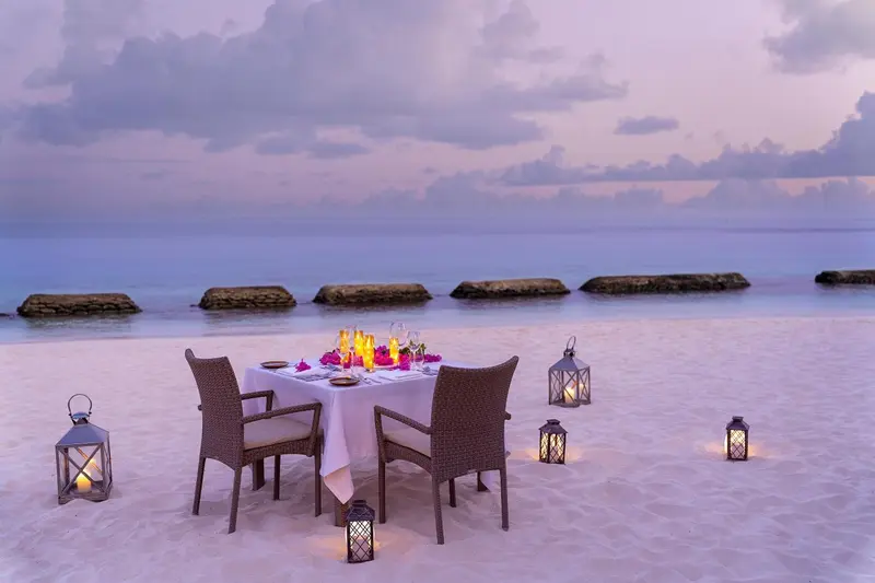 Table dressée pour un dîner romantique sur une plage de sable blanc au coucher du soleil.