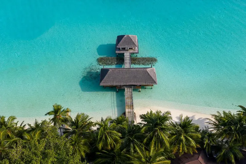 Vue aérienne d'une villa sur pilotis au-dessus d'une eau turquoise et d'une plage de sable blanc.