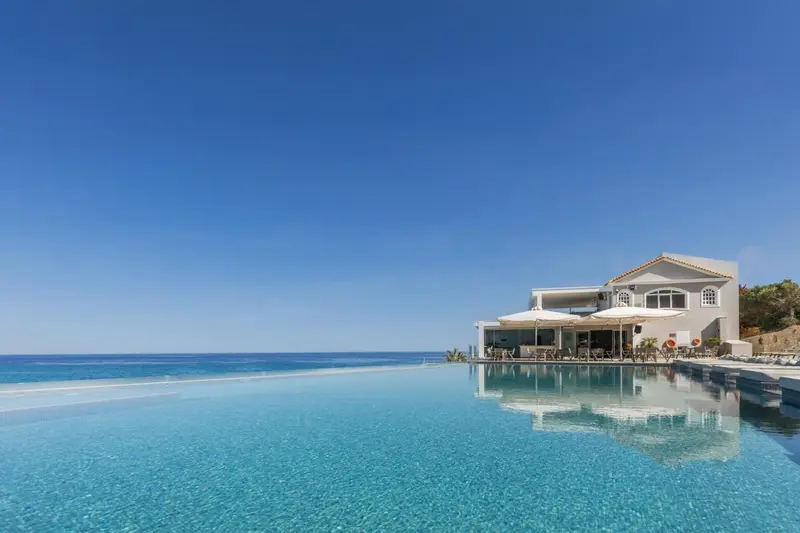 Vue d'une piscine à débordement avec un bâtiment blanc en arrière-plan et la mer au loin.