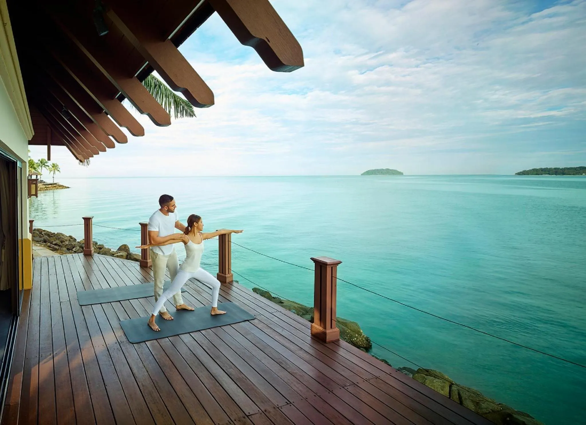 Un couple pratique le yoga sur une terrasse en bois face à la mer.