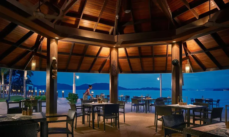 Terrasse ombragée avec tables et chaises donnant sur la mer au crépuscule.