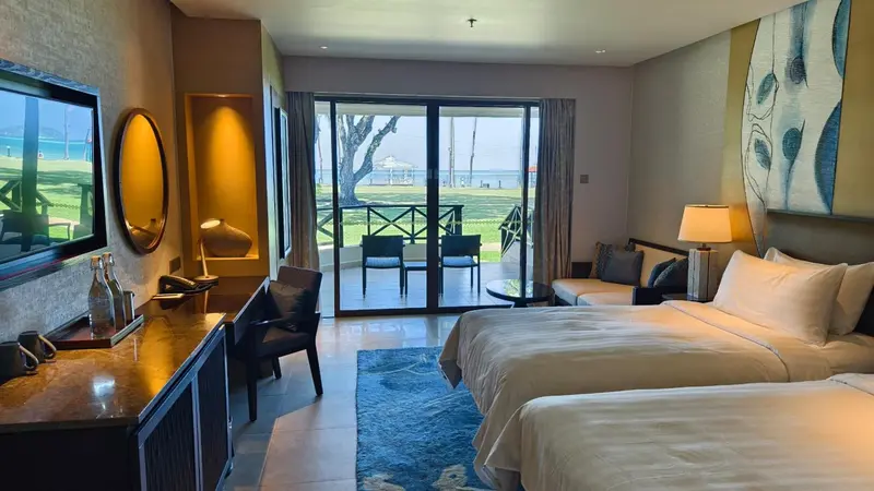 Chambre lumineuse avec un grand lit et un accès à une terrasse donnant sur un jardin et la mer.