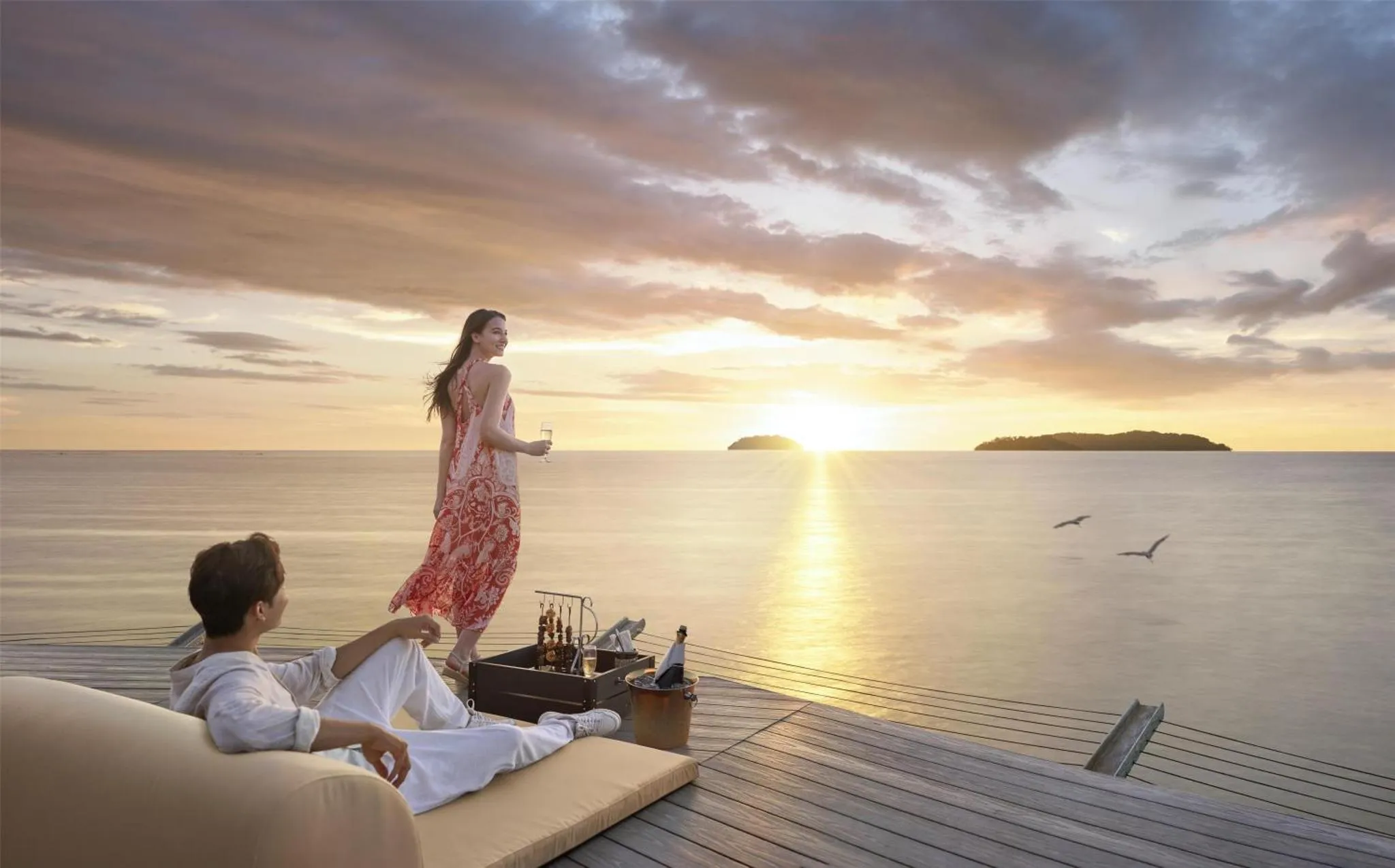 Un couple se détend sur une terrasse en bois face à la mer au coucher du soleil.