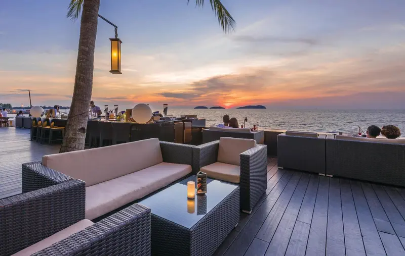 Salon extérieur avec canapés et tables basses face à la mer au coucher du soleil.