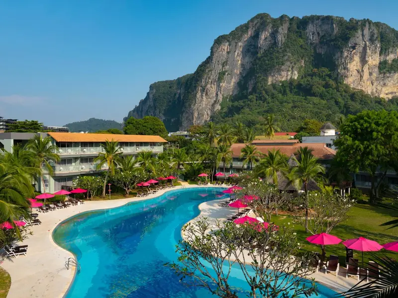Vue aérienne d'une piscine sinueuse bordée de transats roses et de végétation luxuriante, avec une montagne en arrière-plan.