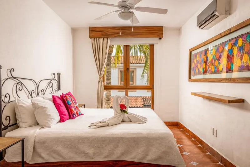 Une chambre lumineuse avec un lit décoré de coussins colorés et une fenêtre donnant sur une cour.