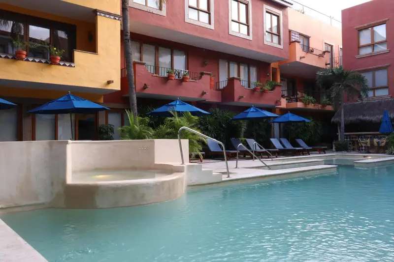 Piscine turquoise bordée de chaises longues et de parasols bleus devant un bâtiment coloré.
