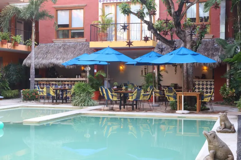 Une piscine turquoise bordée de chaises longues et de parasols bleus, devant un bâtiment coloré.