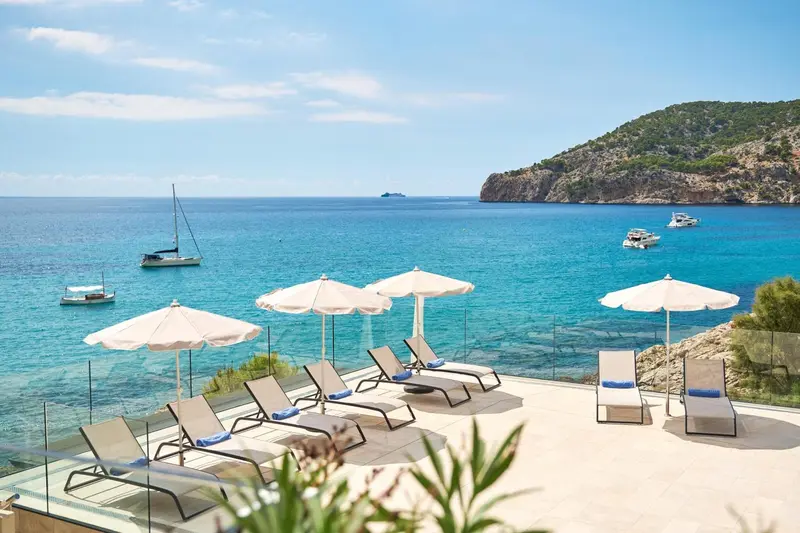 Transats et parasols sur une terrasse surplombant une crique turquoise.