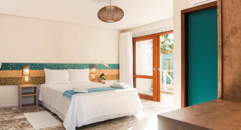 Une chambre lumineuse avec un lit blanc et une porte-fenêtre ouverte sur l'extérieur.