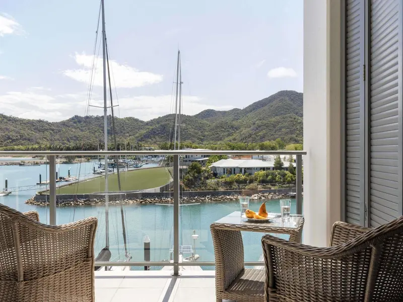 Une terrasse avec une table et des fauteuils offre une vue sur un port de plaisance et des montagnes.