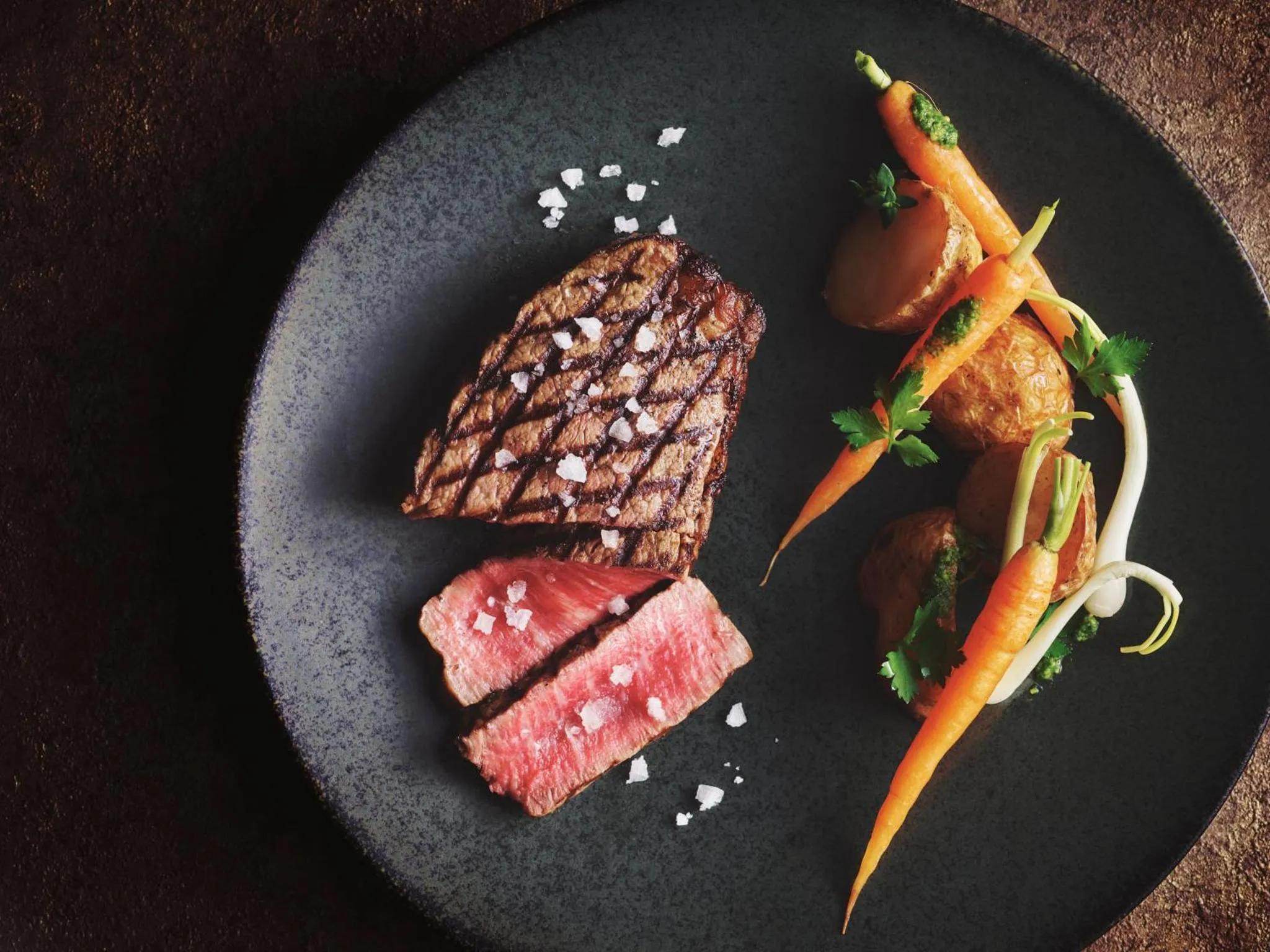 Une portion de viande grillée accompagnée de légumes et d'herbes aromatiques est présentée sur une assiette noire.