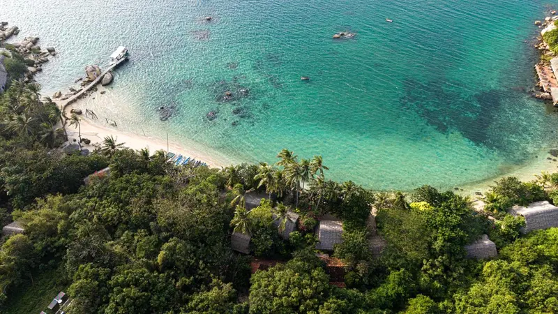 Vue aérienne d'une plage de sable blanc bordée de végétation luxuriante et de bungalows.