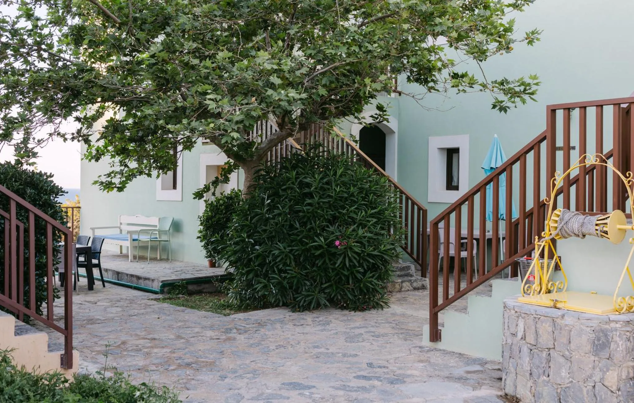 Une cour pavée mène à une terrasse ombragée par un arbre et à un escalier extérieur.