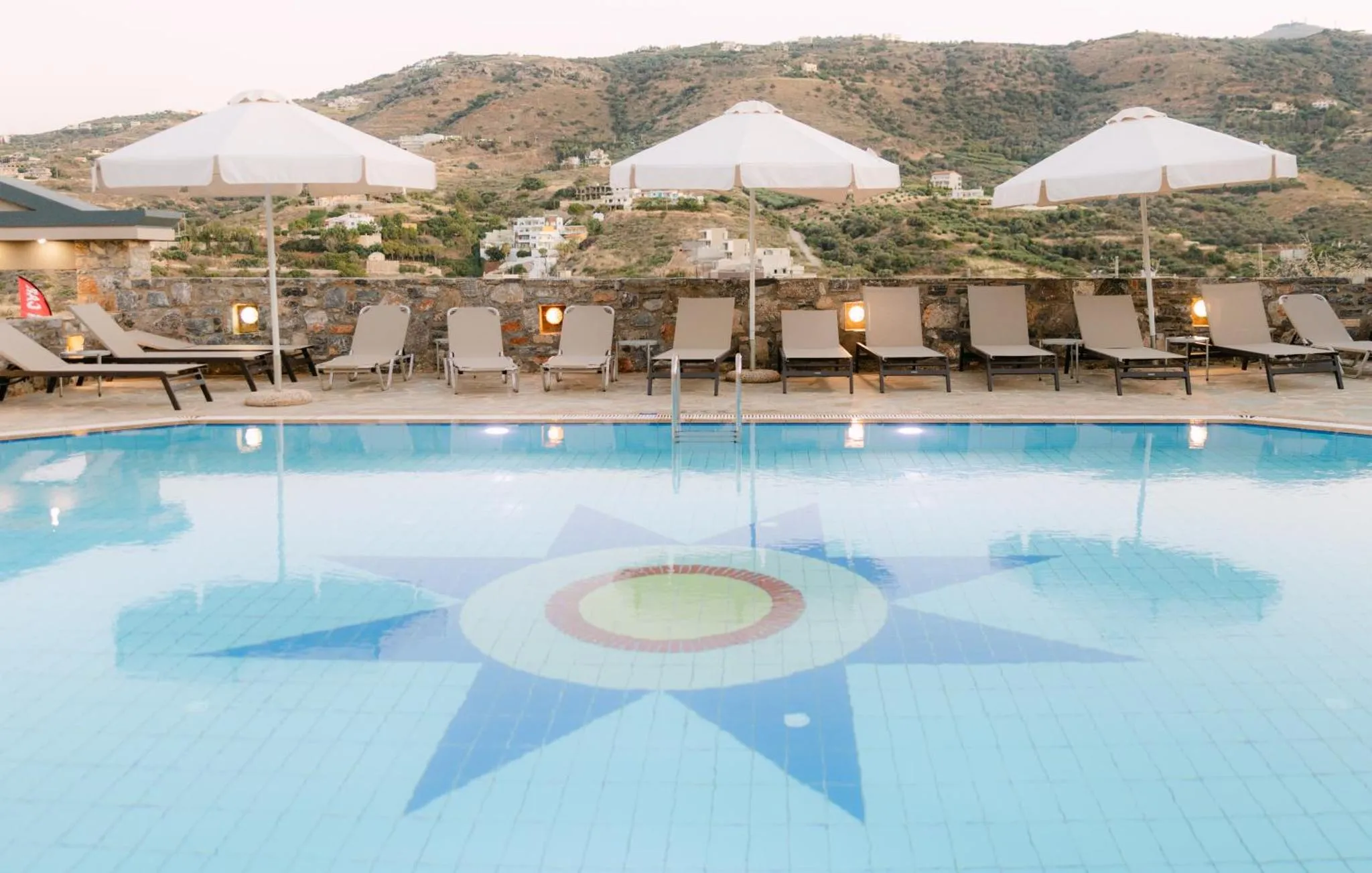 Piscine rectangulaire avec un motif de rosace au fond, entourée de chaises longues et de parasols.
