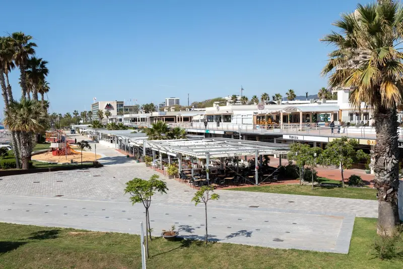 Terrasse ombragée avec tables et chaises en bord de mer, devant des bâtiments.