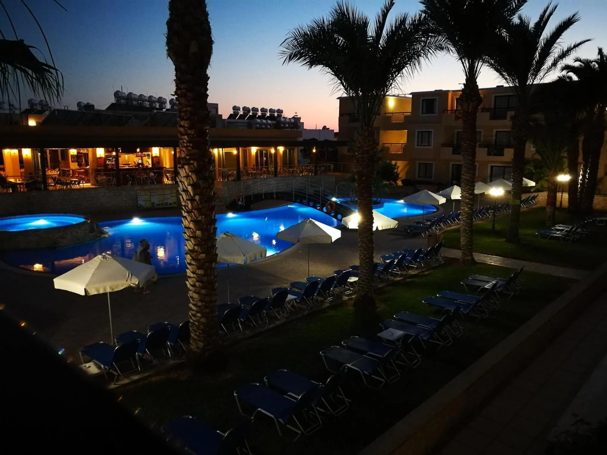 Une piscine illuminée au crépuscule, entourée de transats et de palmiers.