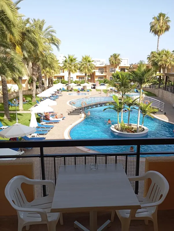 Vue sur une piscine et un jardin depuis un balcon avec table et chaises.