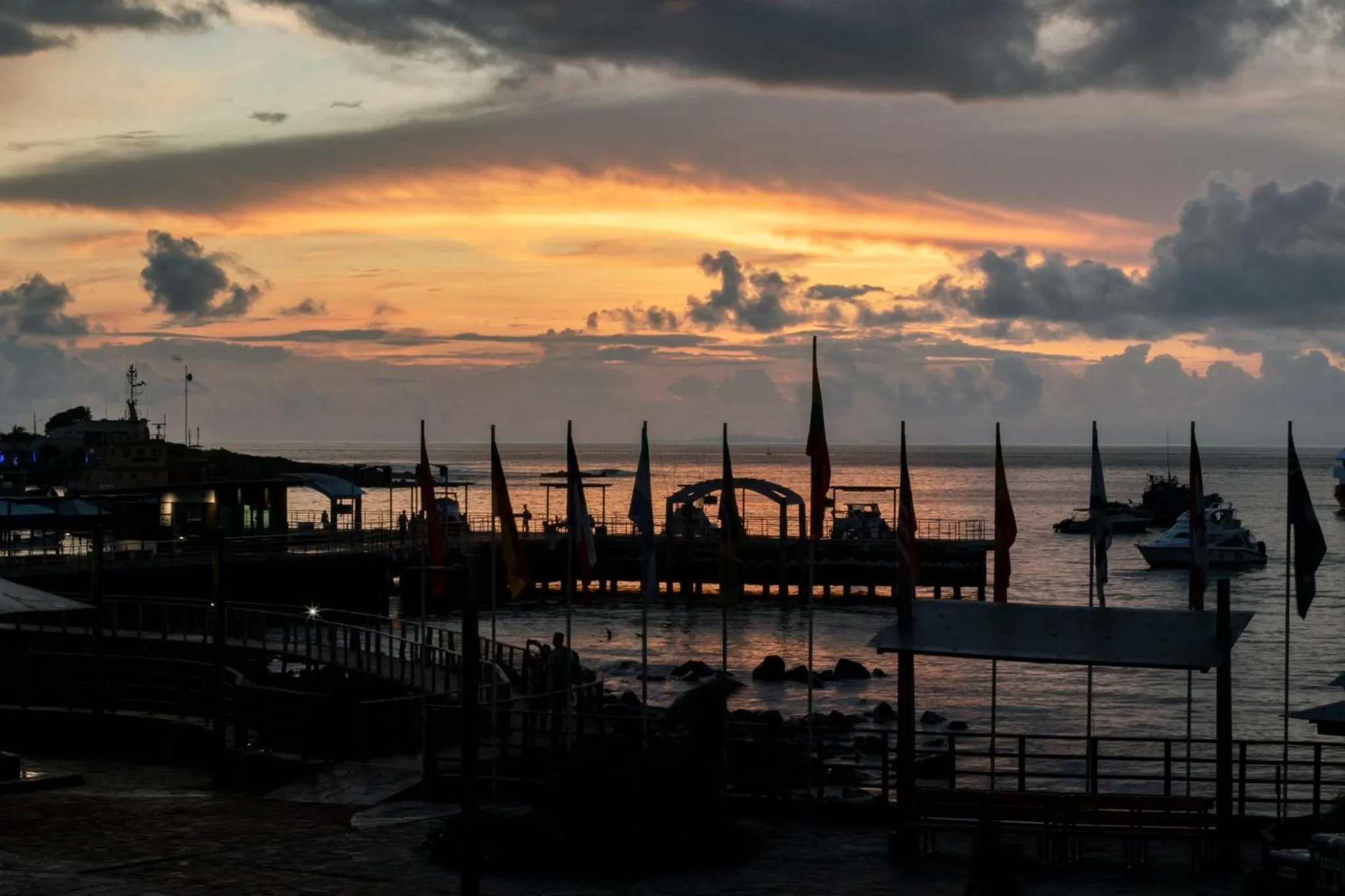 Un ponton s'avance sur l'océan au coucher du soleil, avec des drapeaux et des bateaux amarrés.