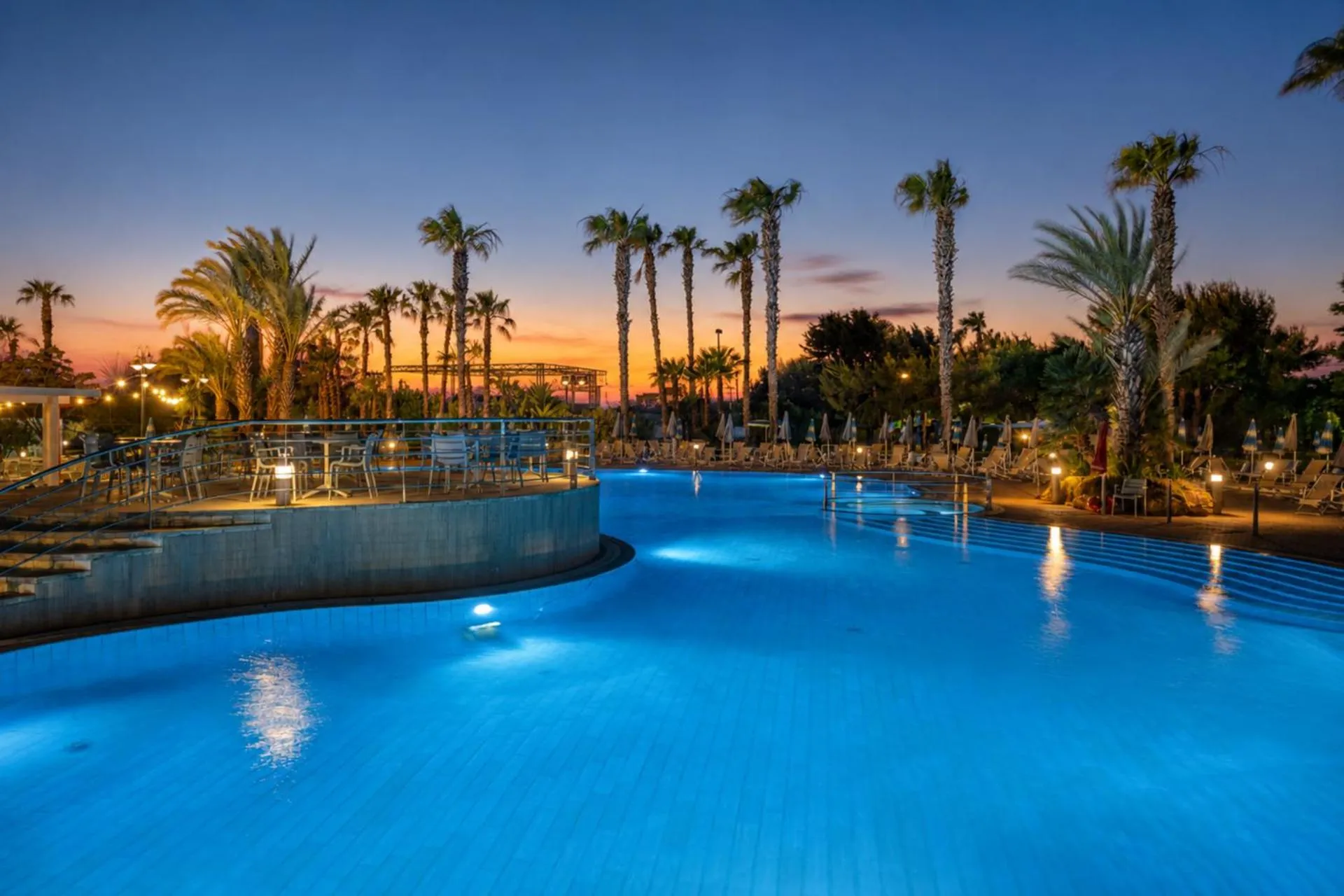 Piscine illuminée au crépuscule avec palmiers et végétation luxuriante.