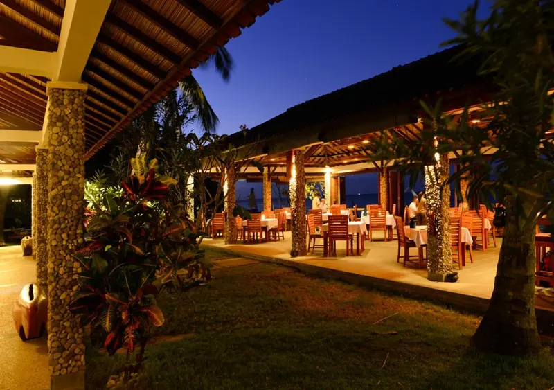 Une salle de restaurant extérieure éclairée avec des tables dressées et une végétation tropicale.