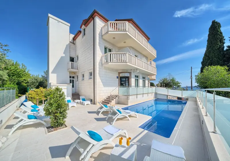 Façade d'un bâtiment blanc avec des transats et une piscine en contrebas.