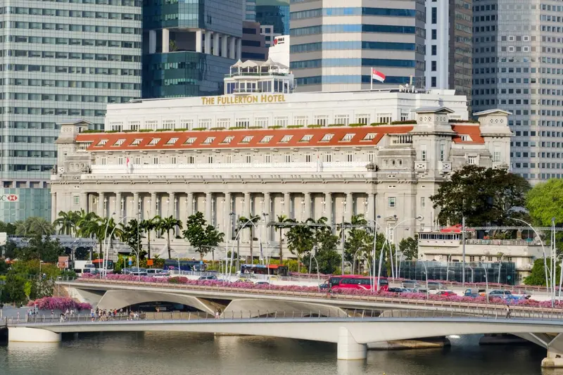 Façade d'un bâtiment colonial blanc avec des balcons et un toit rouge, bordant une rivière et un pont.