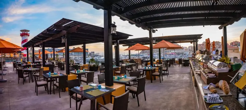 Terrasse ombragée avec tables et chaises donnant sur un port et un phare.