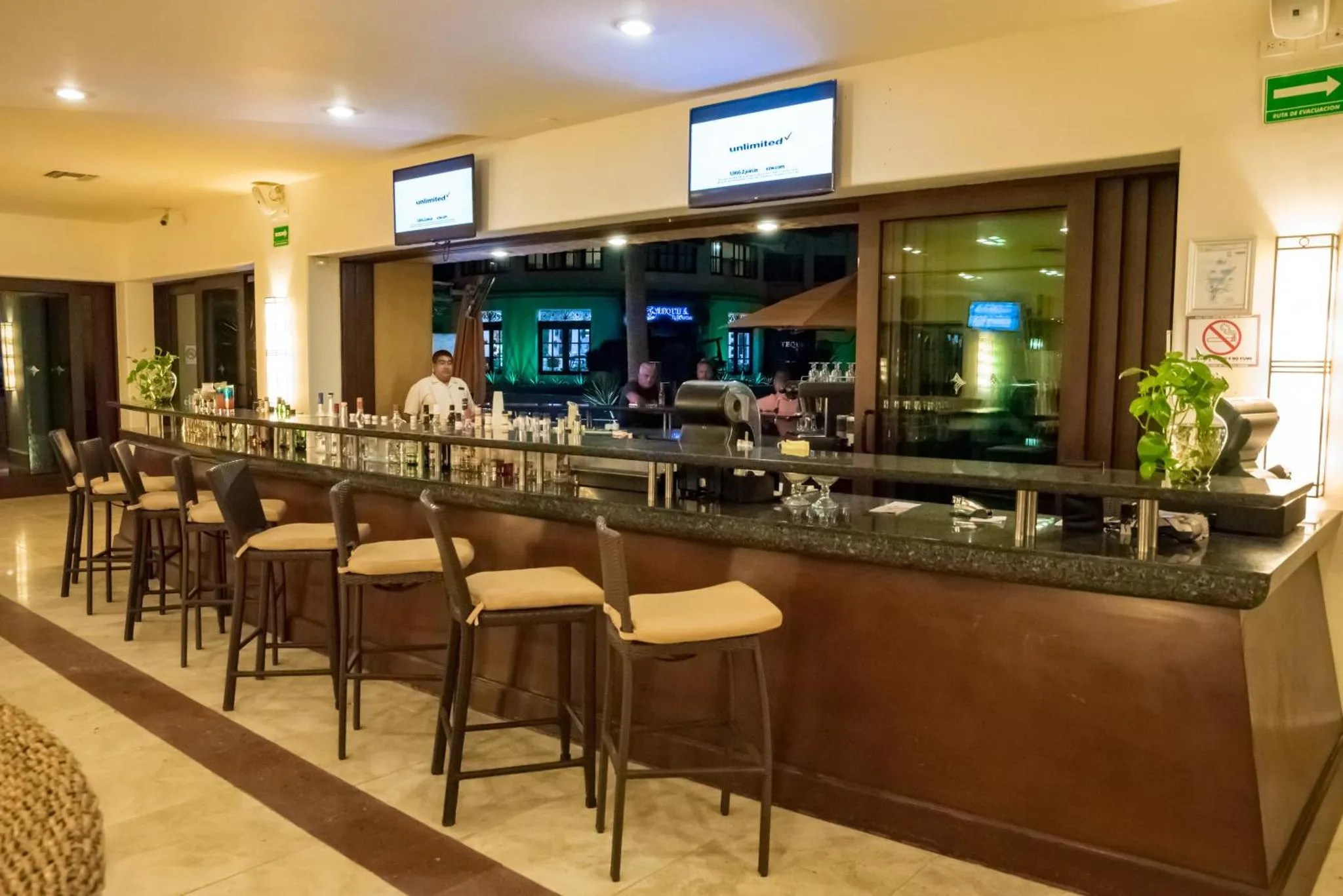 Un long comptoir de bar en bois sombre est aligné de tabourets hauts, avec des bouteilles et des verres.